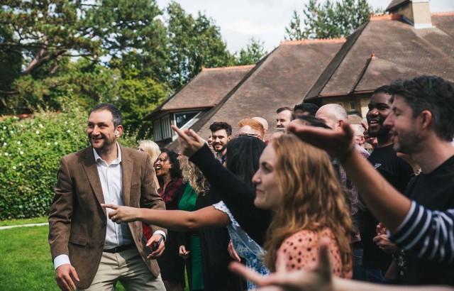 Danny looking up at a photo-taking drone with 20 staff to the right of him raising their hands