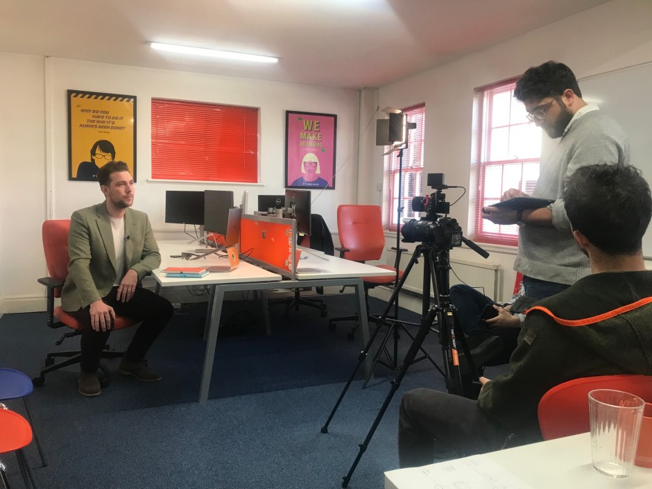 James being filmed in the Elstree office by the Sky crew. He is sitting on a chair facing the cameras, and there are 2 crew members in the foreground.