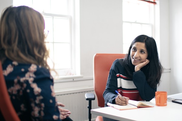 Photo of UX Designer Meera listening to a user whilst writing notes in her orange notebook. Both are sat in orange desk chairs at the Cyber-Duck offices.