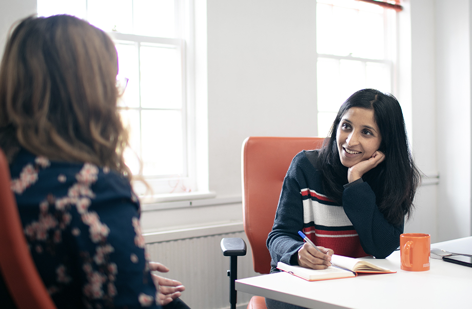 Photo of UX Designer Meera listening to a user whilst writing notes in her orange notebook. Both are sat in orange desk chairs at the Cyber-Duck offices.