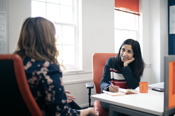 A UX researcher interviewing a user. They are both sitting in office chairs and look like they are in an animated conversation. The user is gesturing with her hands, and the user researcher is smiling and writing her observations down in a notebook.