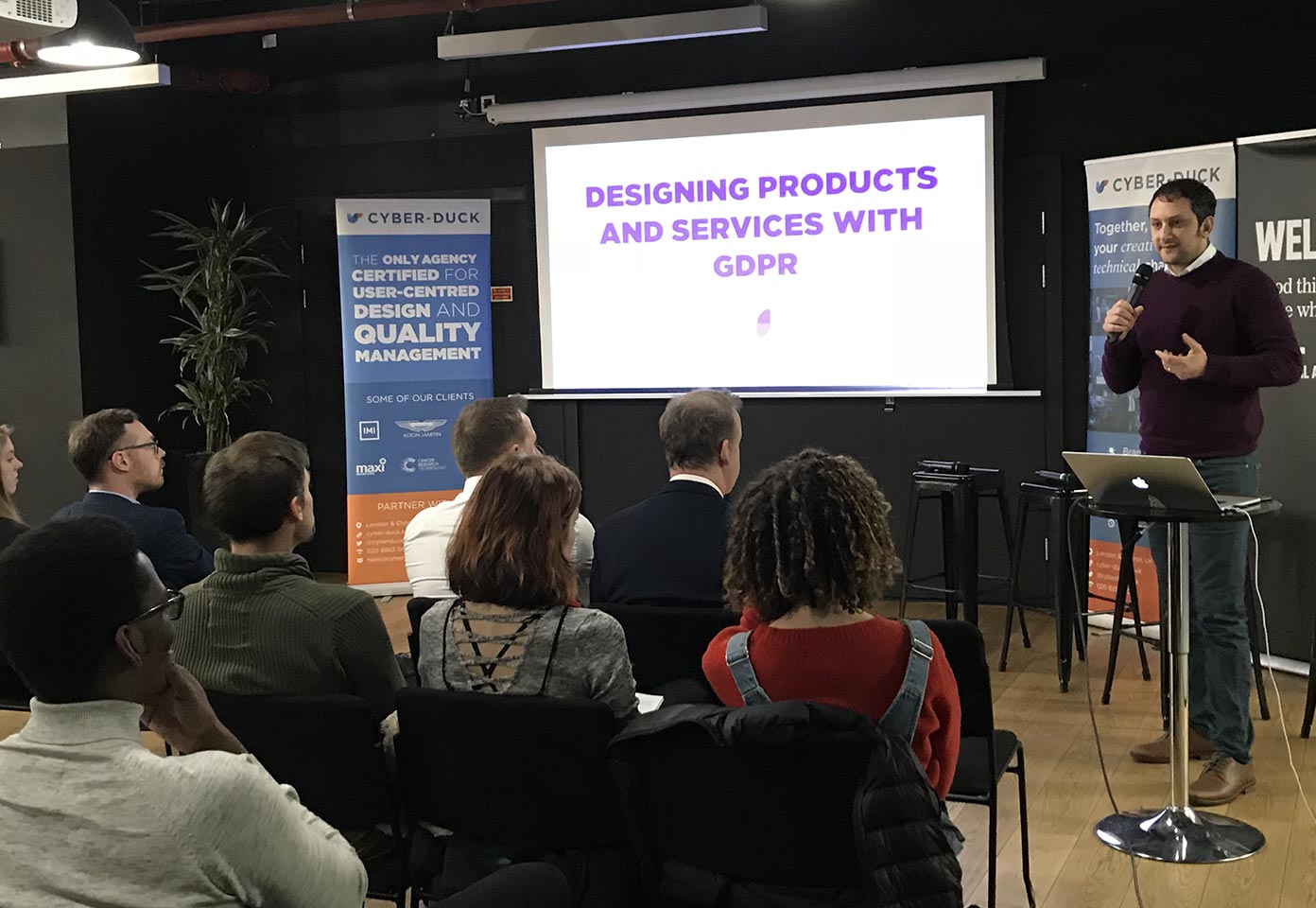 Photo of Cyber-Duck CEO Danny standing in front of an audience with a microphone in his hand. There is a projector showing 'Designing products and services with GDPR' as the title.