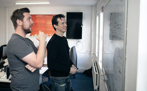 Two man standing in front of white board  talking 