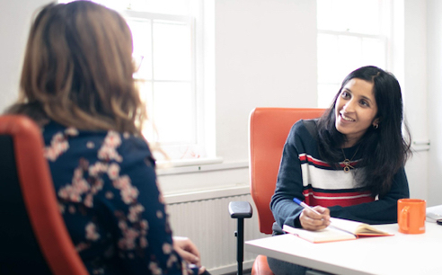 Photo of UX Designer Meera interviewing a user. Both are seated in an office. Meera is writing in a notebook whilst listening to the user with enthusiasm.
