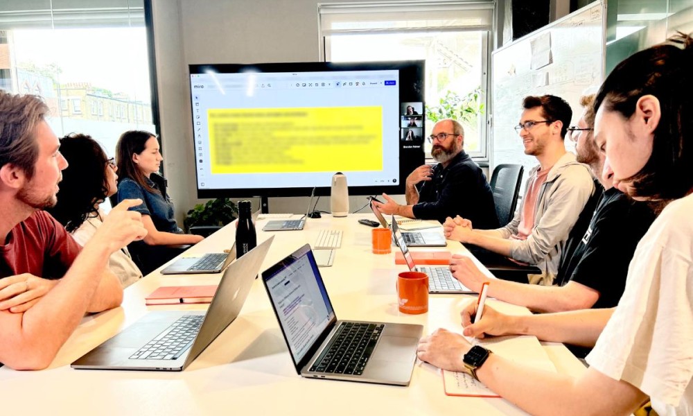 Seven members of the digital product team seated around a conference table discussing a Miro board on a TV screen