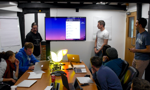 The Cyber-Duck team taking part in a hackathon. Everyone is around a table having animated discussions. There are several laptops and work being shown on the room's TV screen.