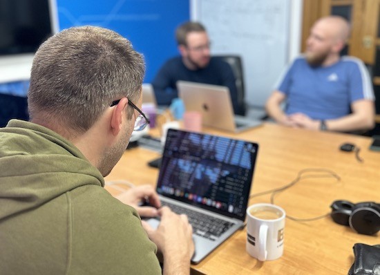 A developer working on an app transformation project, seated at a table in the Cyber-Duck office. Two other developers are having a discussion in the background.