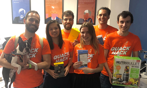 Five of the Cyber-Duck team taking part in a hackathon. They are all smiling at the camera and wearing orange t-shirts that say "Quack Hack". One is holding a cute dog.