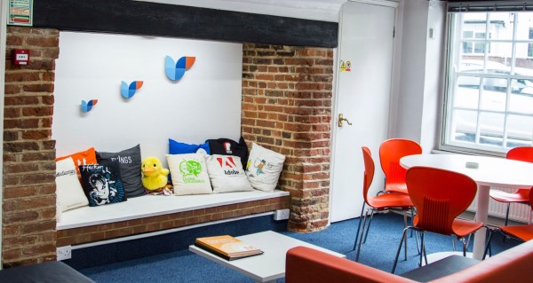 A view of the Elstree office, showing a seating area with cushions, flying ducks on the wall and a table with orange chairs. Sunlight is streaming in through the window.